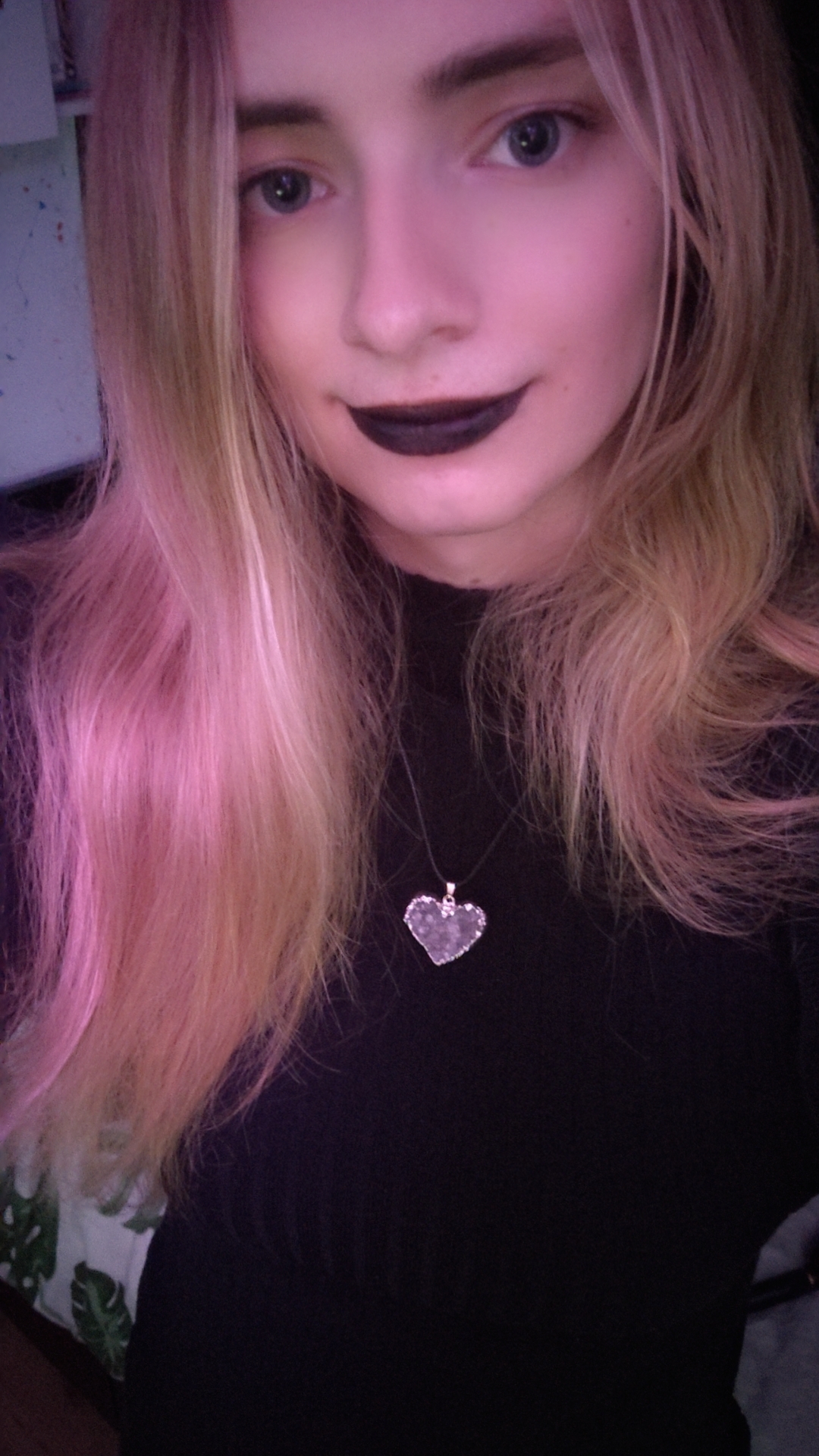 A picture of me, a girl smiling at the camera with blonde hair, blue eyes, wearing black lipstick and a heart-shaped amethyst crystal necklace. I have a long black top on.
